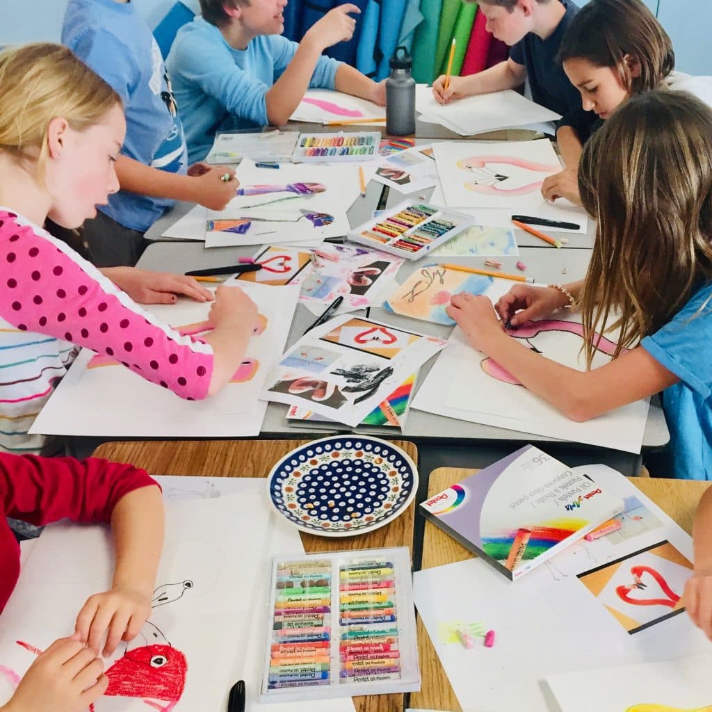 A full table of students drawing with oil pastels, boxes of pastels open between them
