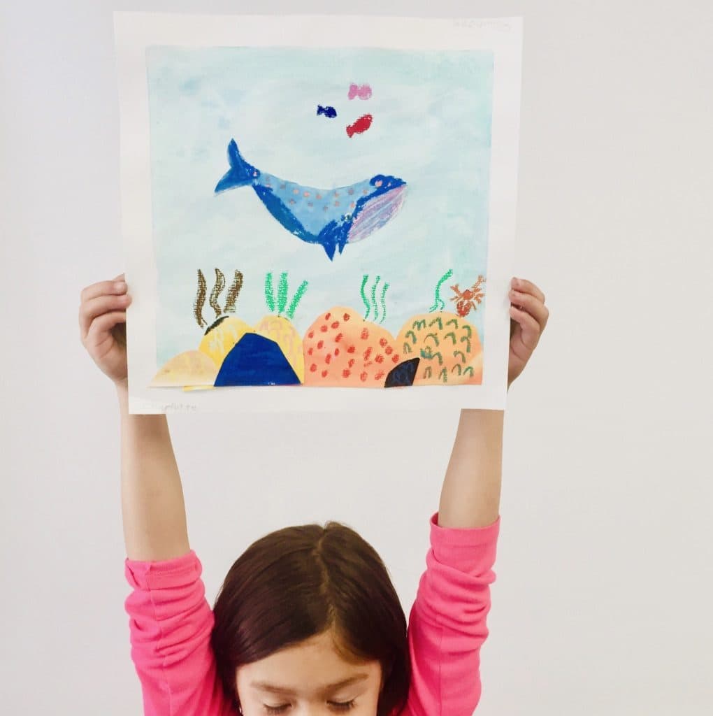 A student holds up an ocean collage: a blue oil-pastel whale over a watercolor sea with paper-cut coral below